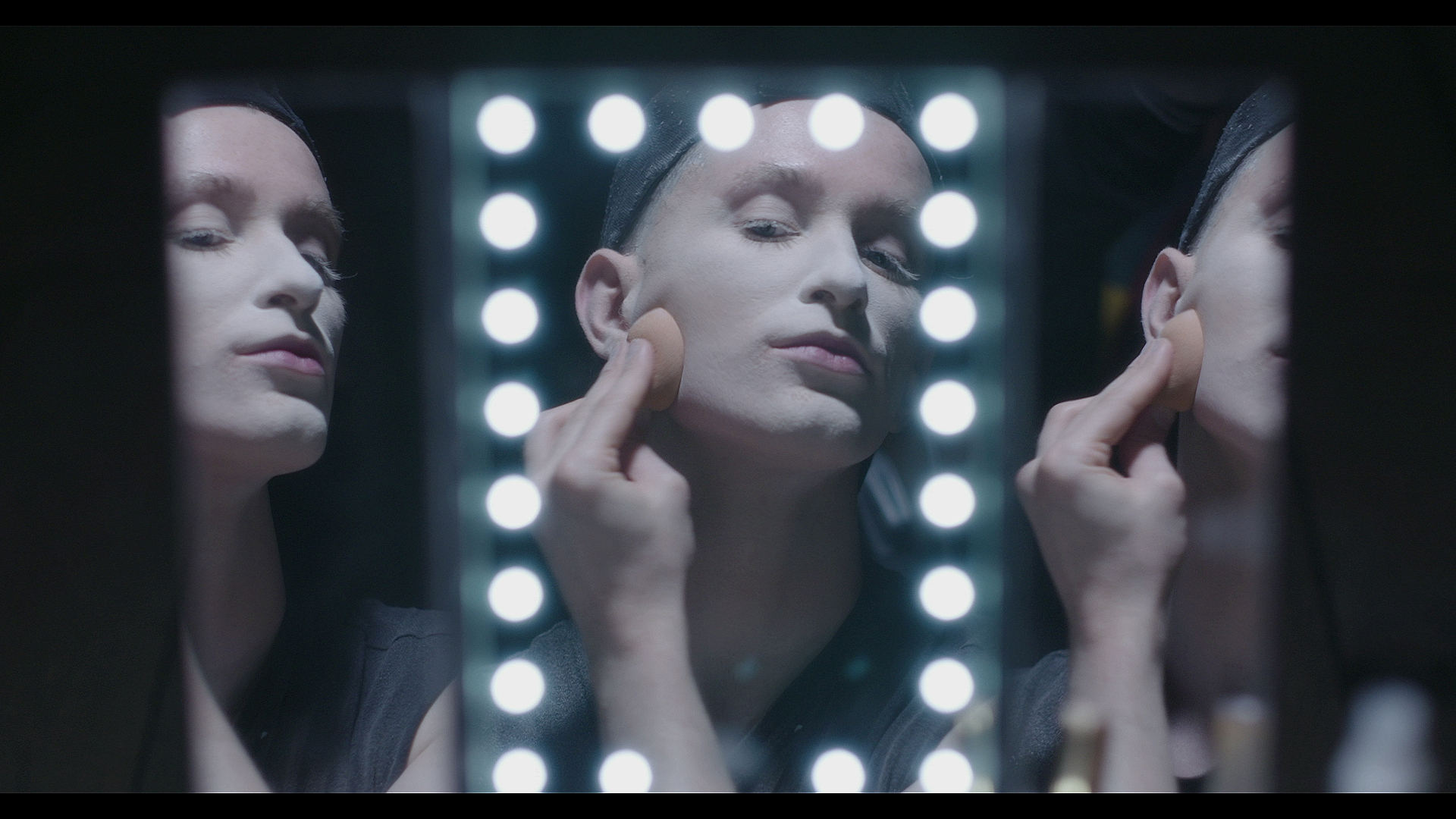 CJ Banks appears looking at herself in three mirrors––one surrounded by lights––powdering white makeup onto her face and wearing a wig cap.