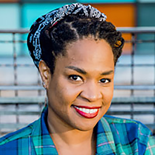 Photo Description: Black woman with medium-brown skin wears a blue and green collared plaid dress and smiles warmly at the camera. She is wearing lipstick and silver hoop earrings. Her hair is in locs and is pulled back under a navy bandana. She is sitting with her hands resting on her lap and arms bent.