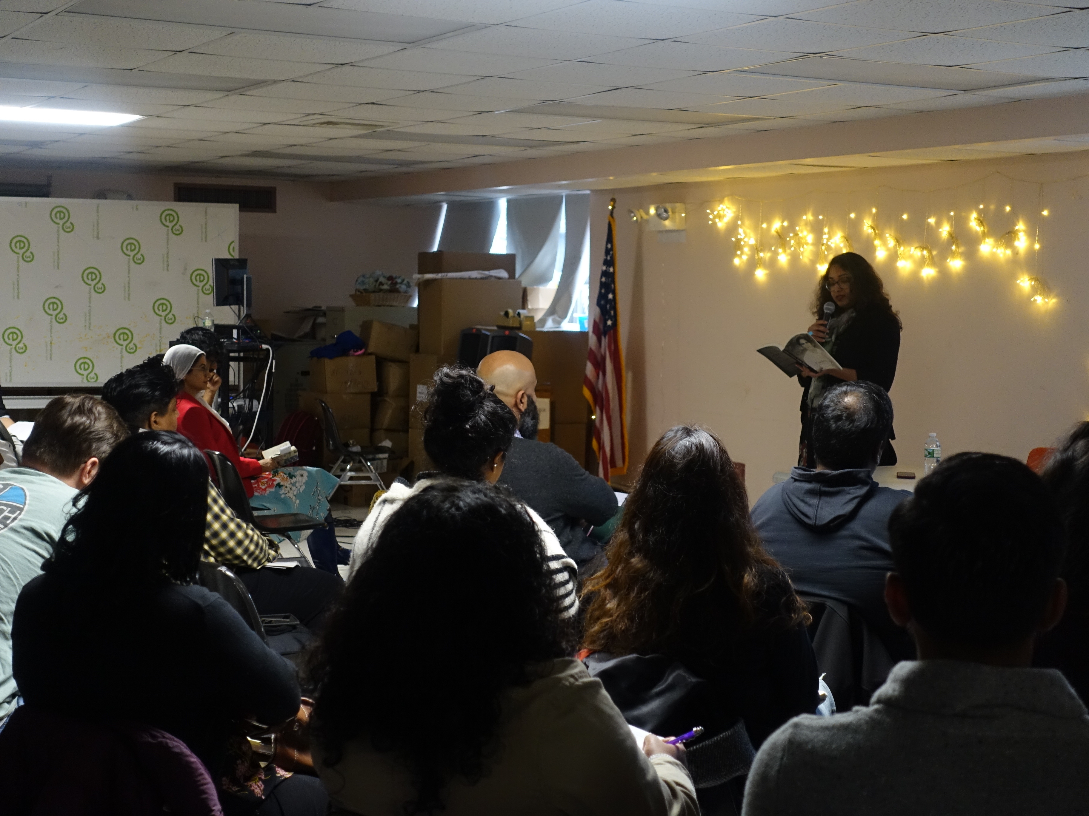 Gaiutra reads from a book, in front of an audience. Behind her is a string of lights.