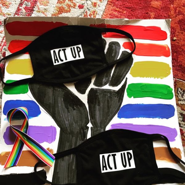 On a hand-painted poster, a painted black fist is raised amid a row of rainbow-colored lines. On top of the poster are two cloth masks with ACT UP logos printed on them, next to a rainbow-colored ribbon.