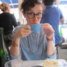 Katie, a curly-haired brunette White person with brown tortoiseshell glasses, a denim button-down and a double-strand necklace, is sipping tea with both hands and looking at the camera.